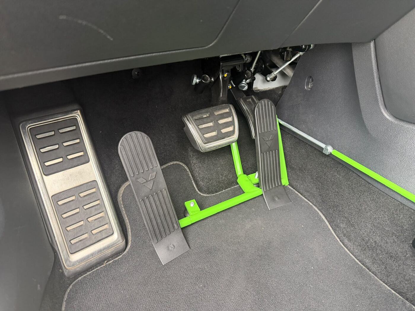 Fußraum eines Fahrschulautos mit Linksgas-Umbau: links neben der
                                  Bremse sitzt ein zusätzliches Gaspedal, das über ein grünes
                                  Gestänge mit dem originalen Gaspedal verbunden ist