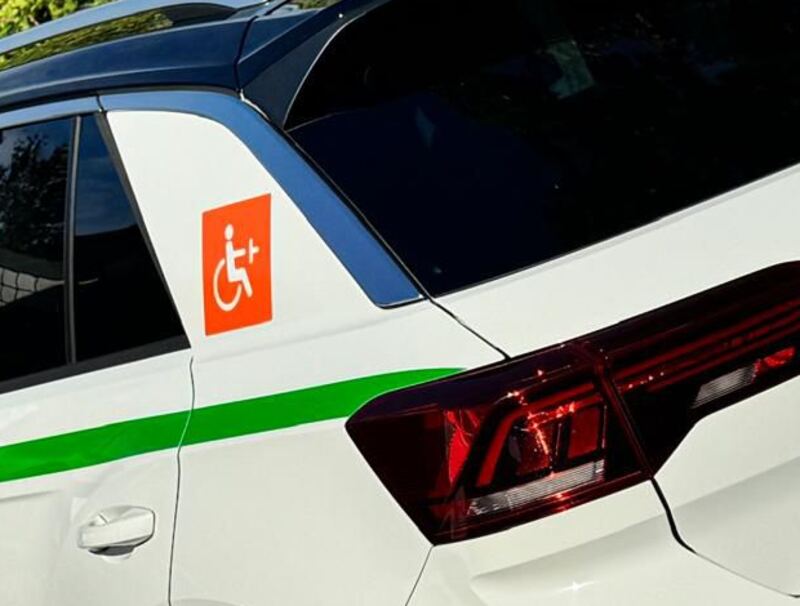 Seite eines weißen Fahrschulautos: ein orangefarbenes
                              Rollstuhlzeichen auf der hinteren Scheibe, darunter ein grüner
                              Streifen über der Tür