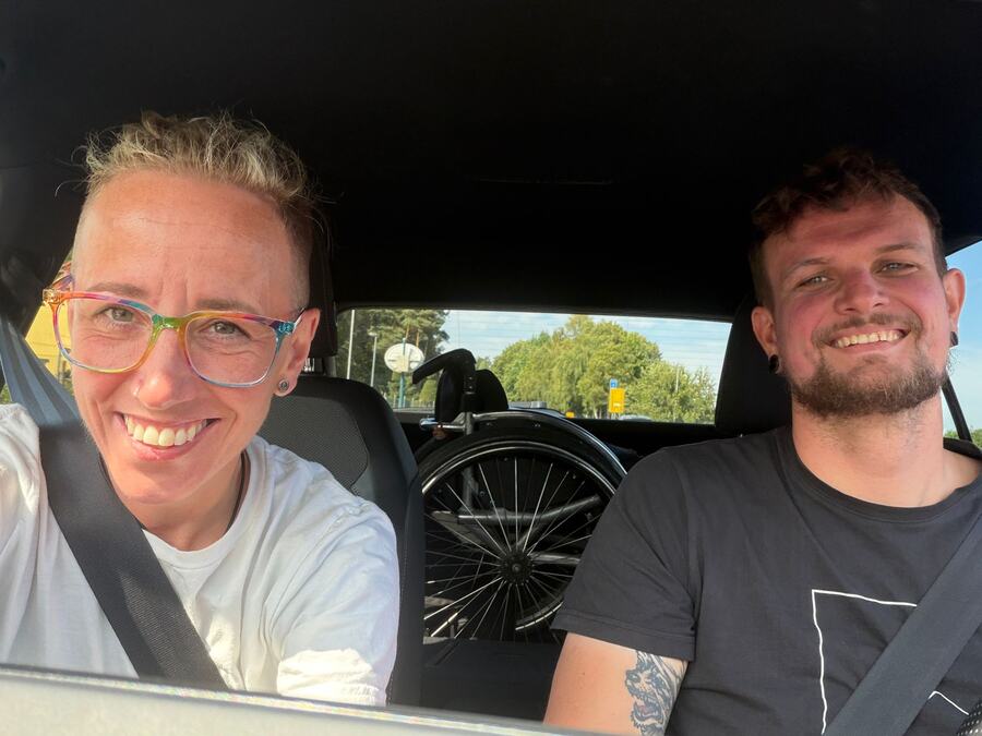 Sarah und ein Fahrschüler angeschnallt im Auto, hinter
                                  ihnen liegt ein zusammengeklappter Rollstuhl
