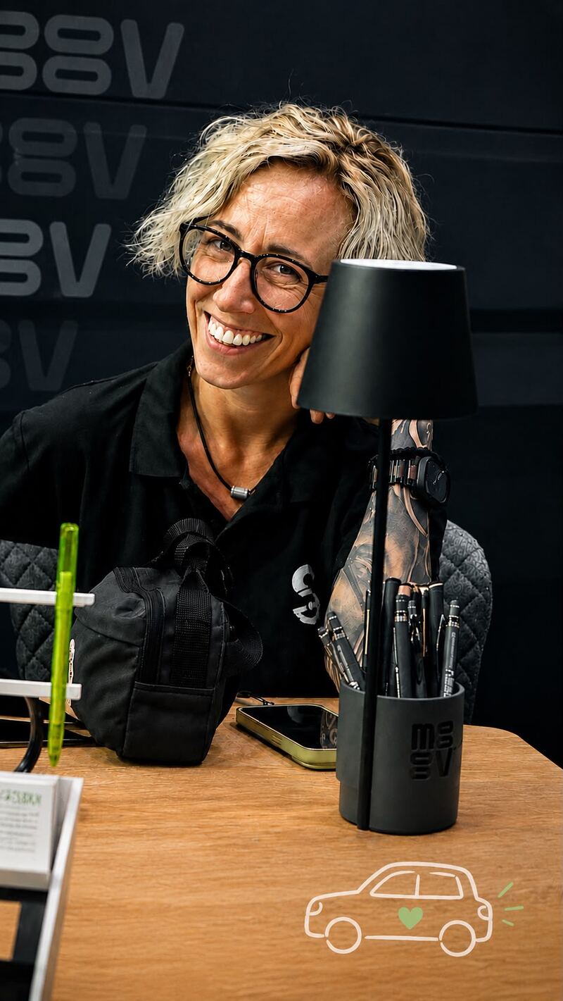 Sarah sitzt lächelnd an einem Tisch, den Kopf in die Hand
                              gestützt, daneben eine Tischlampe und ein Becher mit Stiften