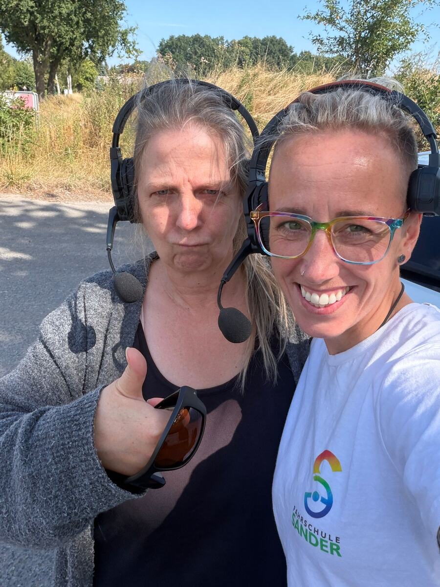 Sarah und eine Kollegin am Straßenrand, beide mit
                                  Headset; die Kollegin zeigt den Daumen nach oben